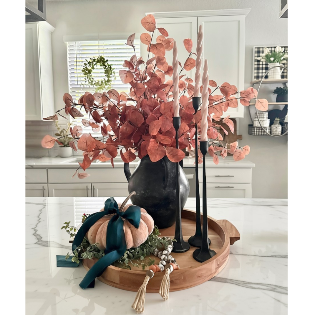 Autumn-themed tabletop centerpiece in a real kitchen setting, featuring a black pitcher vase filled with coppery eucalyptus branches, a small peach pumpkin with a dark teal bow, black candlesticks with white taper candles, and a round wooden tray on a white marble counter.