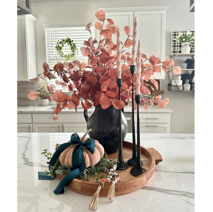 Autumn-themed tabletop centerpiece in a real kitchen setting, featuring a black pitcher vase filled with coppery eucalyptus branches, a small peach pumpkin with a dark teal bow, black candlesticks with white taper candles, and a round wooden tray on a white marble counter.
