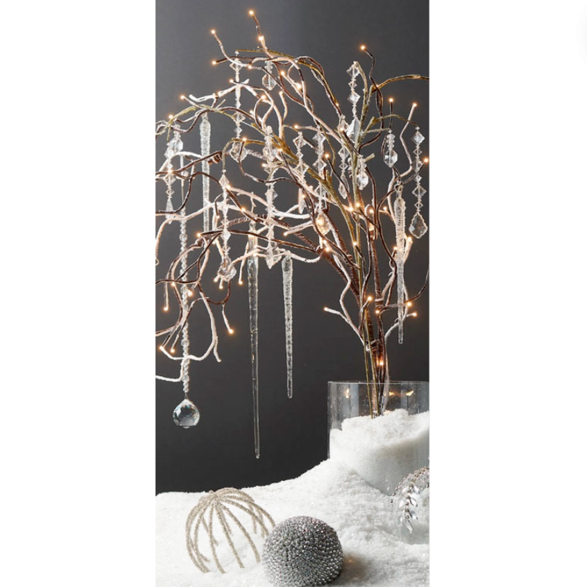A decorative lighted branch with brown branches and hanging silver and white lights, placed over a glass vase filled with snow, set against a dark background.
