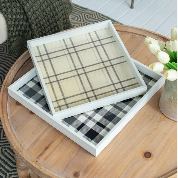 Two square decorative trays with white frames are stacked on a round wooden table in a styled living room setting. The top tray features a beige and brown plaid pattern, while the bottom tray has a black and white buffalo plaid pattern.