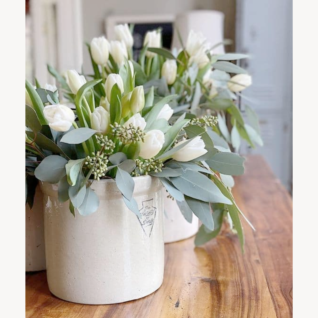 A bundle of white real touch tulips arranged in a ceramic vase, with greenery and paper bows, placed on a wooden surface.