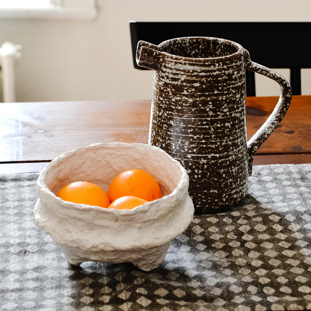 Ceramic pitcher and a white paper maché footed bowl with oranges on a patterned tablecloth