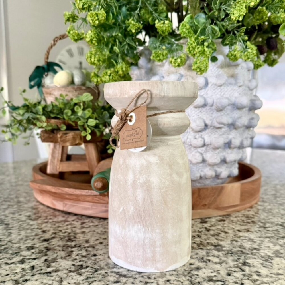Decorative mango wood candle holder with a tag on a countertop with plants and a wooden stool in the background.