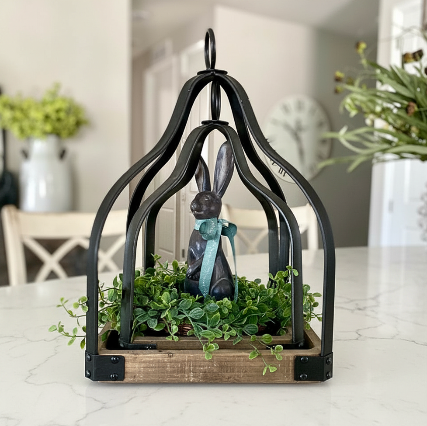 A decorative black metal lantern set with a rustic wood base, holding green trailing foliage and a dark rabbit figurine wearing a light blue bow, set on a white marble countertop in a softly blurred home interior.