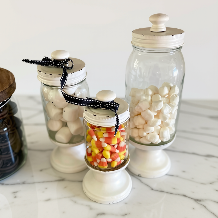 Three clear glass canisters with white pedestal bases and white lids are arranged on a white marble surface. The canisters are filled with white candies, candy corn, and dark brown items, and two of them have black-and-white polka dot ribbon bows on the lids.