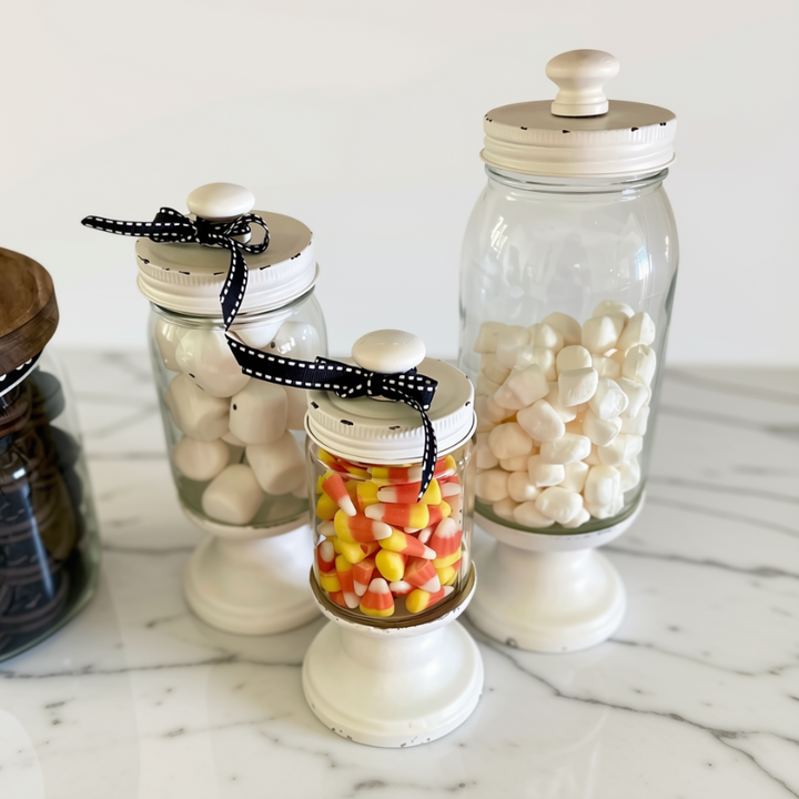 Three clear glass canisters with white pedestal bases and white lids are arranged on a white marble surface. The canisters are filled with white candies, candy corn, and dark brown items, and two of them have black-and-white polka dot ribbon bows on the lids.