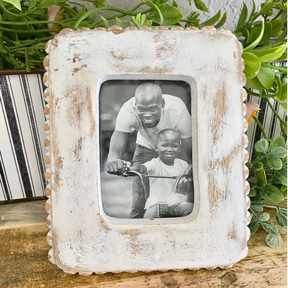 White distressed picture frame with a photo of a man and child on a bike, placed on a wooden surface with plants in the background.