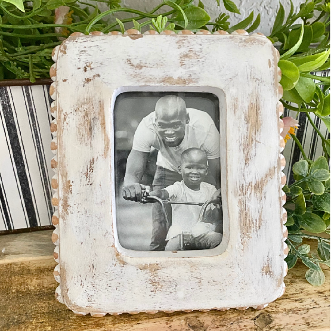 White distressed picture frame with a photo of a man and child on a bike, placed on a wooden surface with plants in the background.