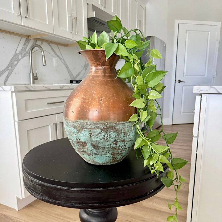 A large decorative vase with a distressed copper upper half and a distressed turquoise lower half, filled with trailing green ivy, displayed on a round black side table in a bright kitchen setting.