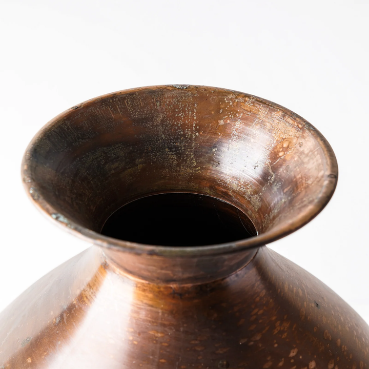 Close-up of a rustic copper-toned metal pot or vessel showing the flared rim and dark interior against a plain white background.