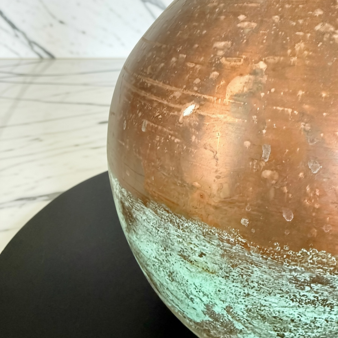 Close-up of a decorative metal vase with a distressed copper and turquoise patina finish, resting on a black circular base.