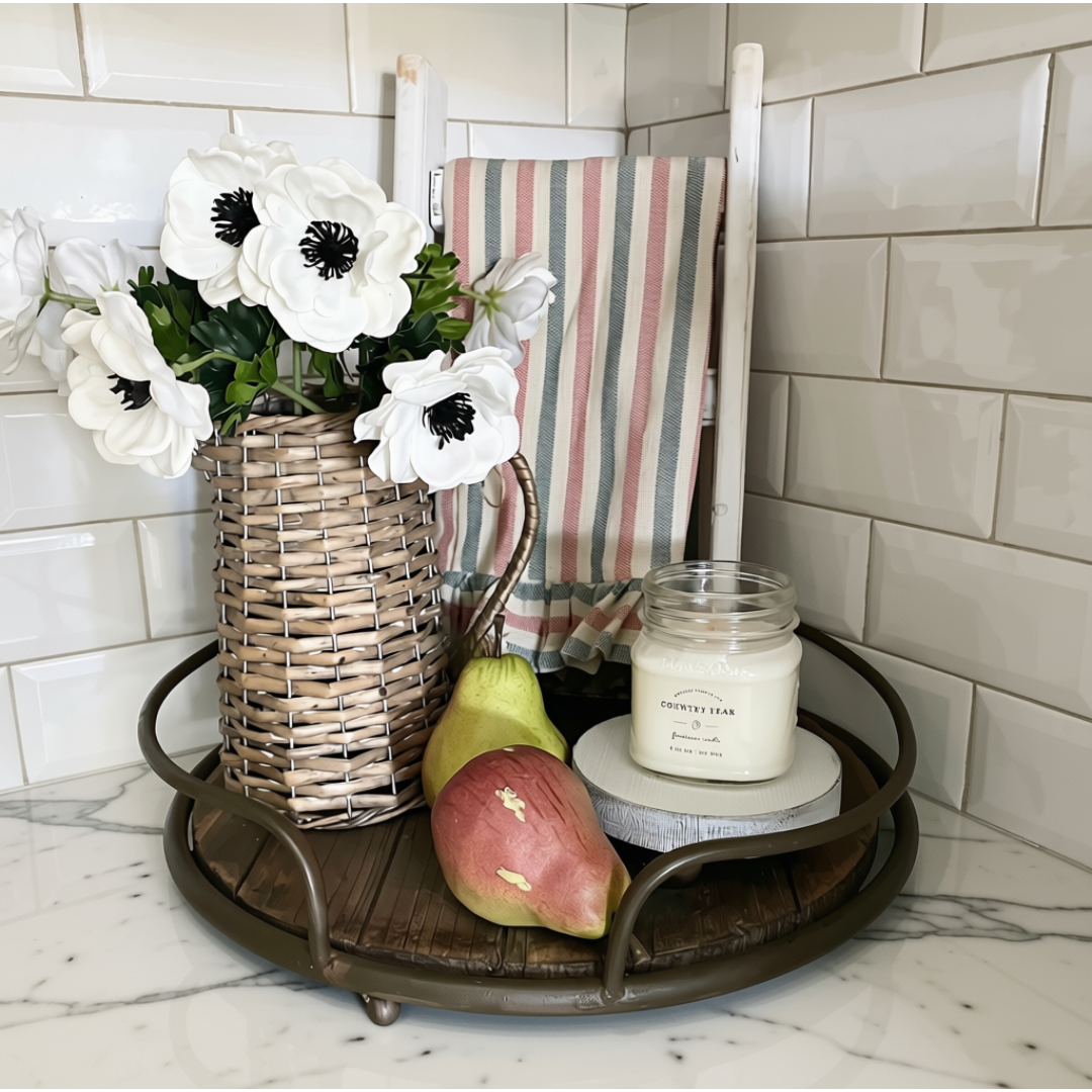 A round dark metal tray with a wooden slatted top is styled in a kitchen corner with white subway tile and a marble countertop. The tray holds a wicker basket vase with white anemone flowers, a striped cloth, a pear, a red apple, and a glass jar candle labeled 'Country Pear.