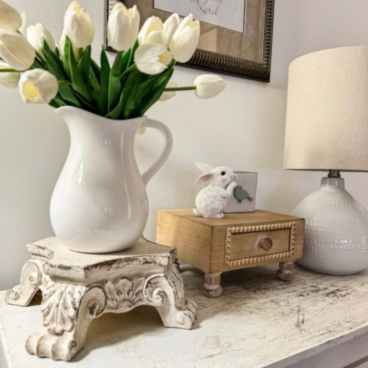 A white ceramic pitcher filled with white tulips sits on a distressed white pedestal stand on a tabletop. A small wooden drawer with a white rabbit figurine on top and a white table lamp are visible in the softly lit room setting.