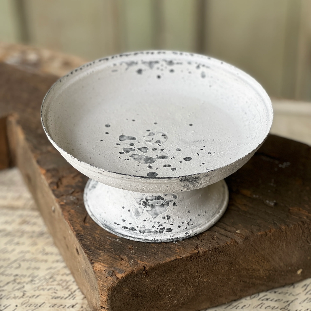 A small white pedestal plate with a distressed, speckled finish, displayed on a rustic wooden block.