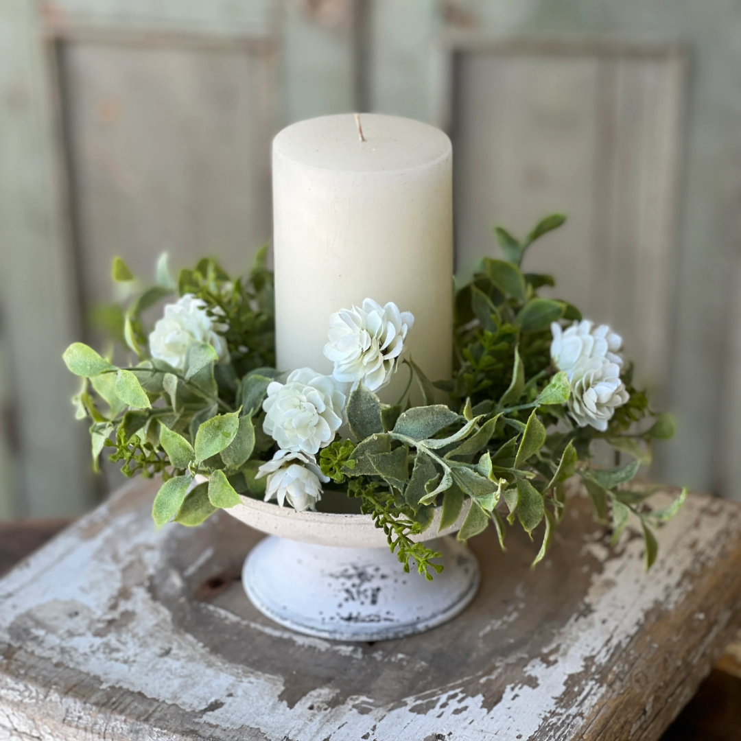A white pillar candle displayed in a distressed white pedestal holder, surrounded by green eucalyptus leaves and small white flowers, set on a rustic wooden surface with a blurred background.