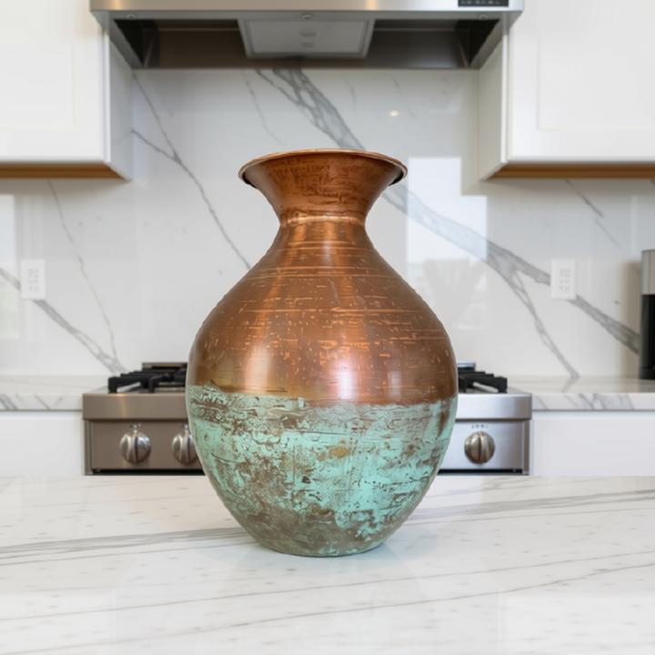 A large decorative vase with a distressed copper upper half and a patina-finished green lower half, displayed on a white marble kitchen countertop with a marble backsplash and stainless steel stove in the background.