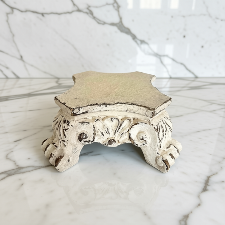 A small decorative pedestal riser with a distressed finish, featuring a hexagonal top and an ornate carved base with claw-like feet, shown on a white marble surface.