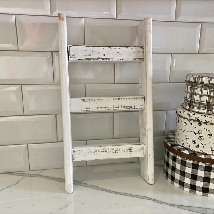 A small rustic countertop wooden ladder shelf with a distressed white finish, standing on a marble countertop against a white subway tile backsplash. Decorative stacked boxes are visible on the right side.