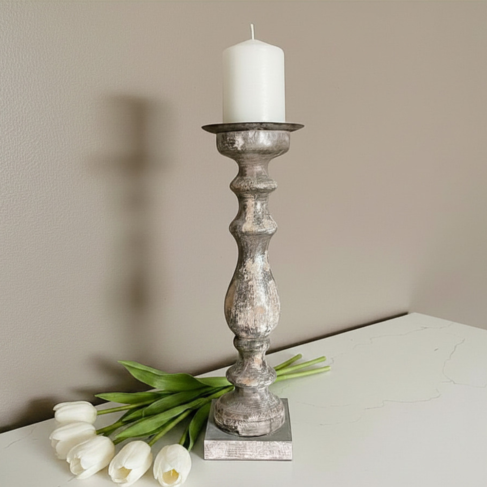A distressed wood candlestick holder with a white pillar candle, styled on a white tabletop next to white tulips against a beige wall.