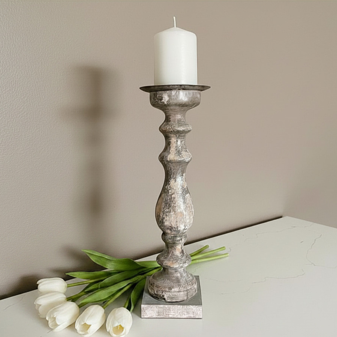 A distressed wood candlestick holder with a white pillar candle, styled on a white tabletop next to white tulips against a beige wall.