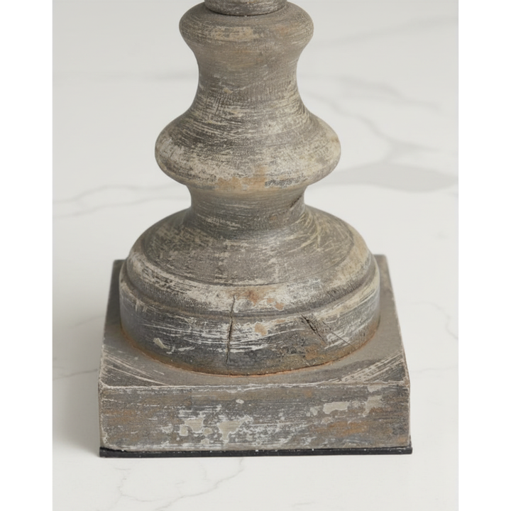 A close-up of a distressed gray wooden pedestal or candlestick base with a square plinth foundation, set against a light marble background.