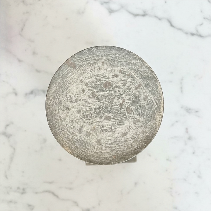 A round, distressed silver metal plate with a heavily scratched and weathered finish, photographed from directly above on a white marble surface.