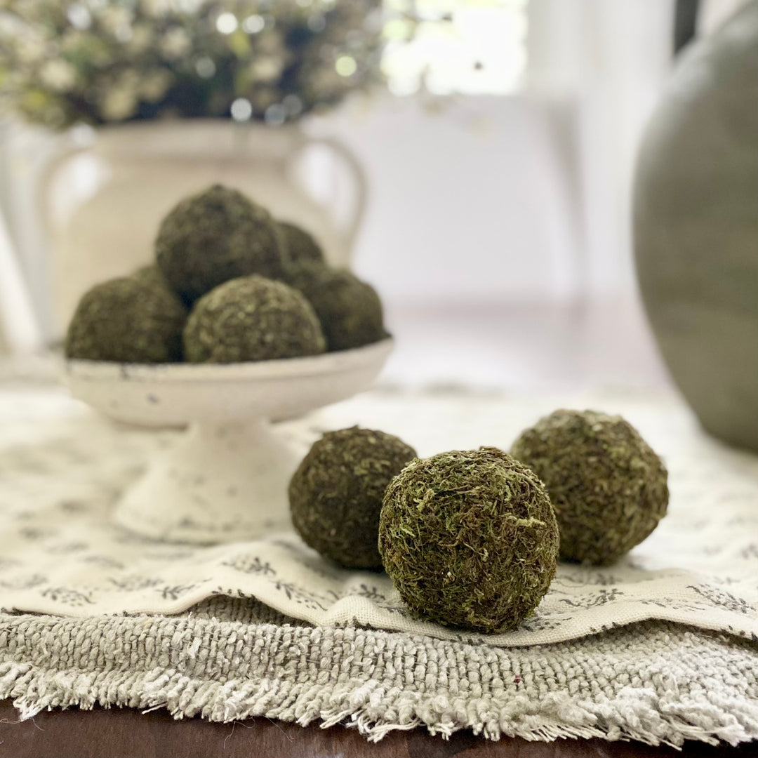 A box containing nine dried moss balls displayed on a white pedestal plate with a rustic design, placed on a table with decorative items in the background.