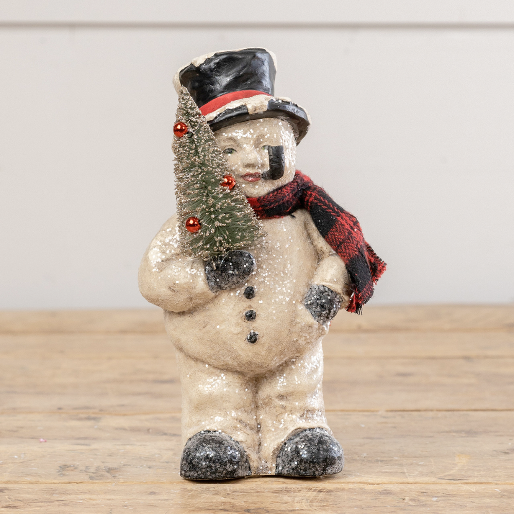 Decorative snowman figurine holding a small Christmas tree on a wooden surface.