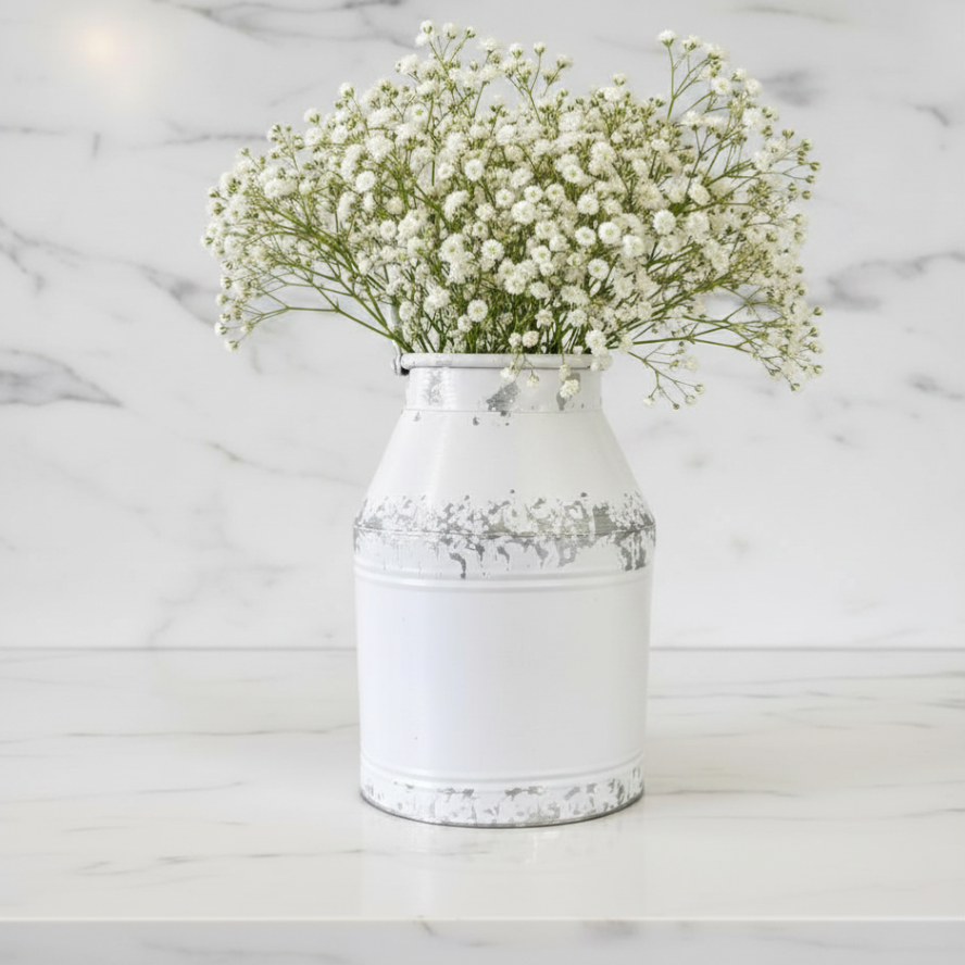 A white distressed metal milk-can style vase filled with white baby's breath flowers, set on a white marble surface against a white marble background.
