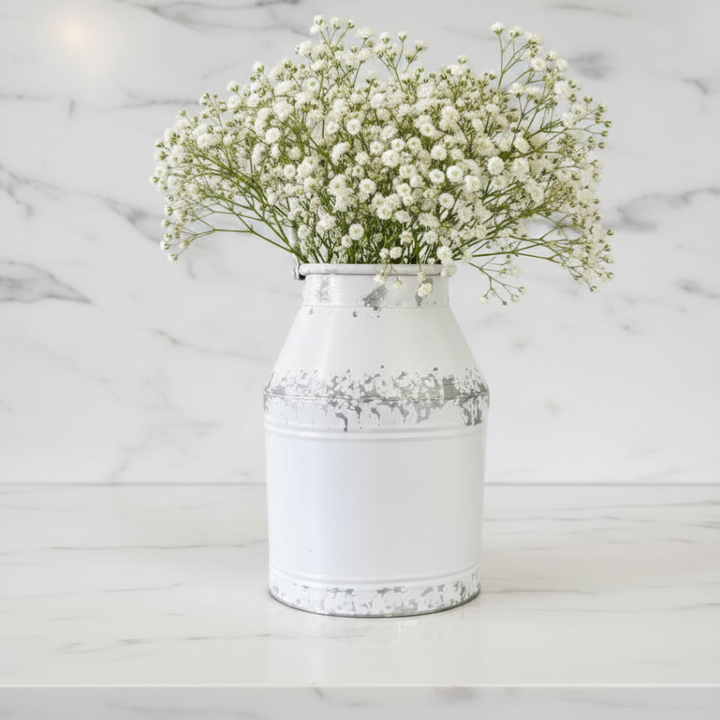 A white distressed metal milk-can style vase filled with white baby's breath flowers, set on a white marble surface against a white marble background.