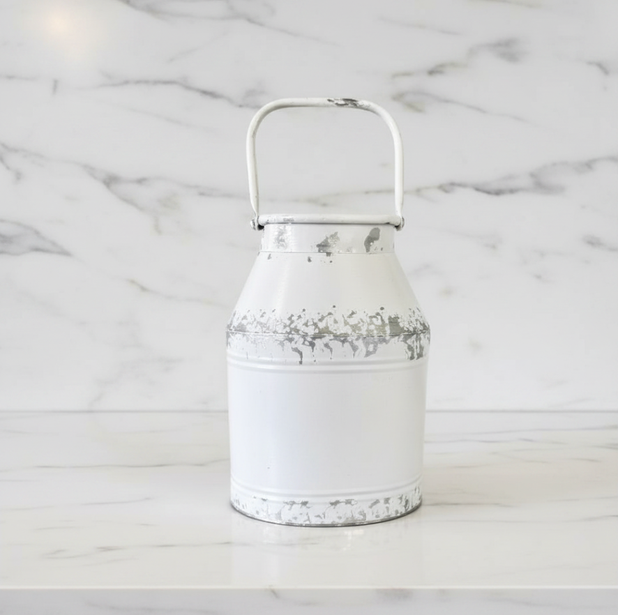 A white distressed metal milk can with a top handle, set on a white marble surface against a matching marble background.
