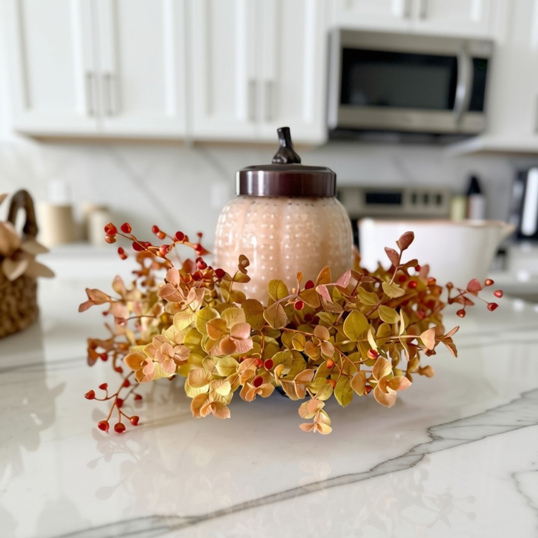 A decorative autumn centerpiece with a textured beige jar, a dark lid, and a surrounding wreath of faux yellow, peach, and orange leaves with small red berries, displayed on a white marble kitchen countertop.