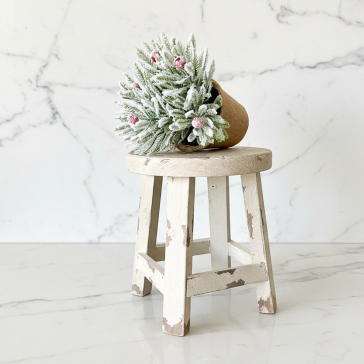 A rustic distressed white wooden stool with a round seat, holding a small frosted green pine arrangement in a brown pot, set against a white marble background.
