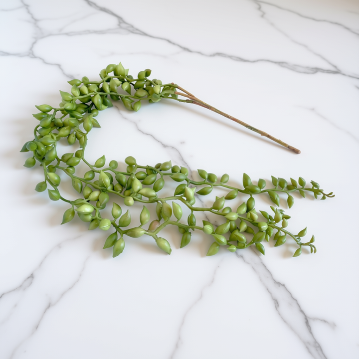 Green artificial trailing vine branch with small oval leaves and brown stems, arranged on a white marble surface.