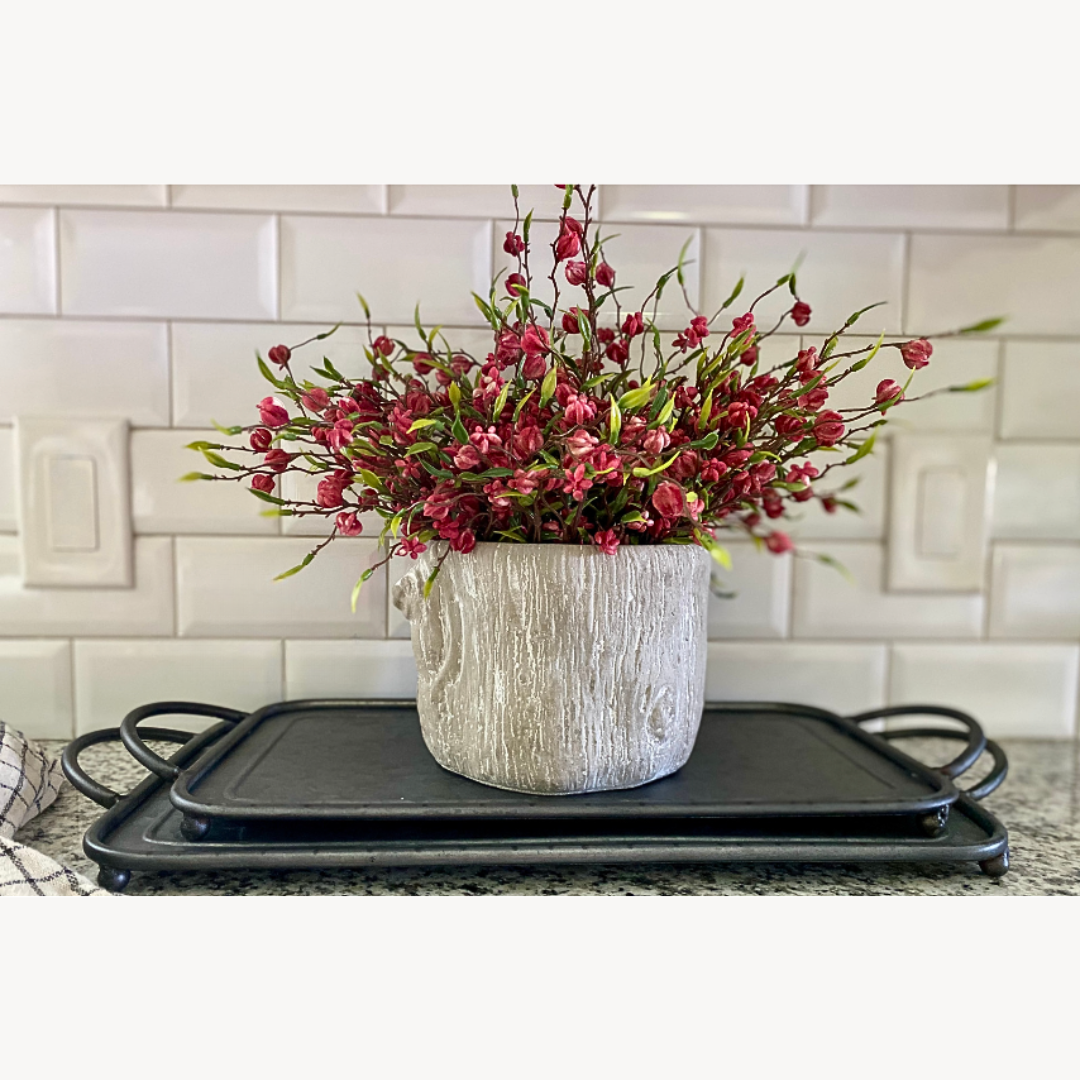 Decorative flower arrangement in a textured faux tree bark pot on a black tray against a tiled wall.