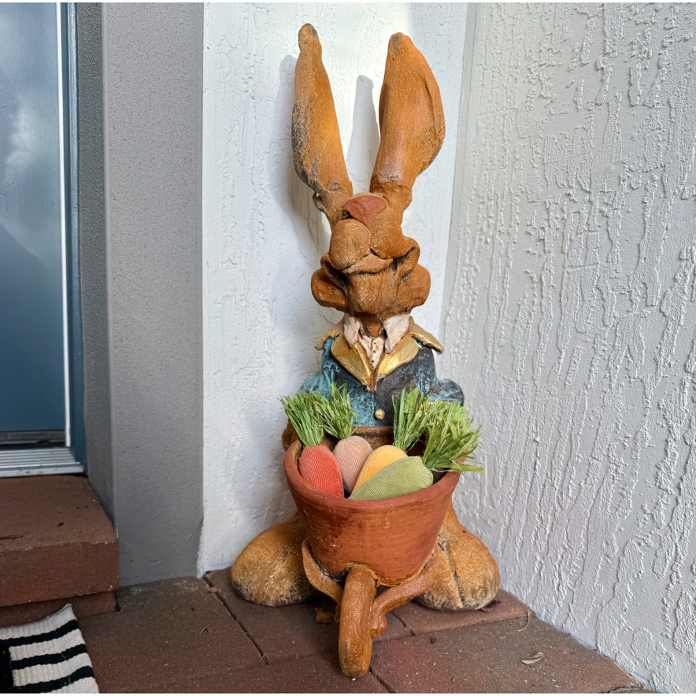 Decorative rabbit statue with a basket of fabric carrots against a white wall.
