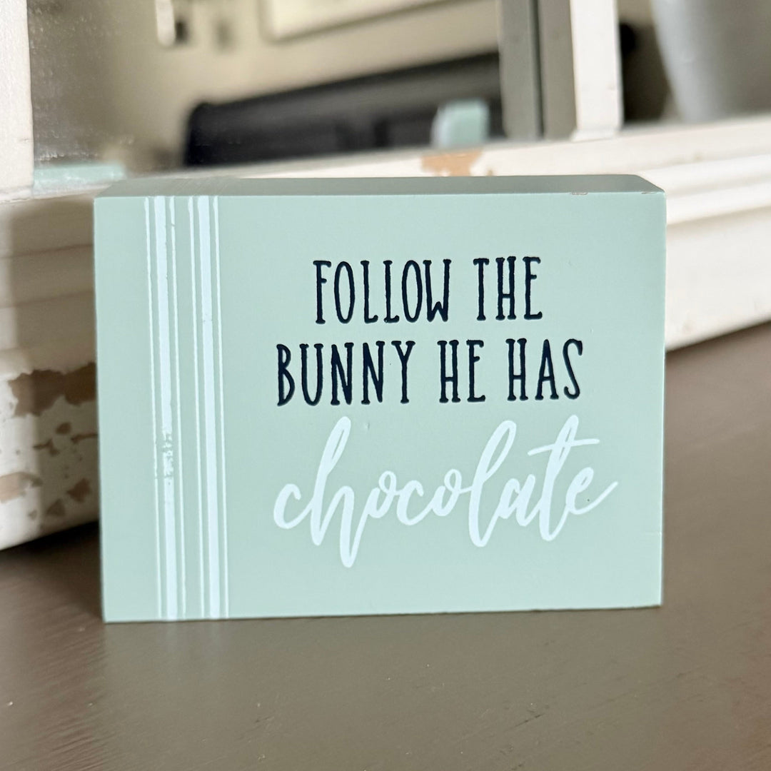 Light blue sign with text 'Follow the bunny he has chocolate' on a surface.
