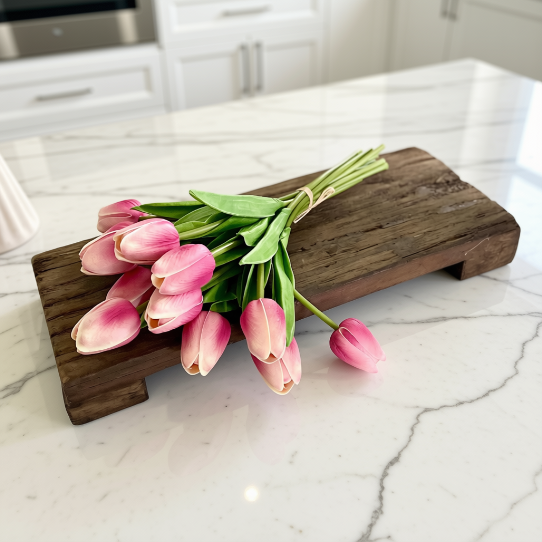 A bouquet of pink tulips with green stems rests on a rustic dark wood decorative riser placed on a white marble kitchen countertop.