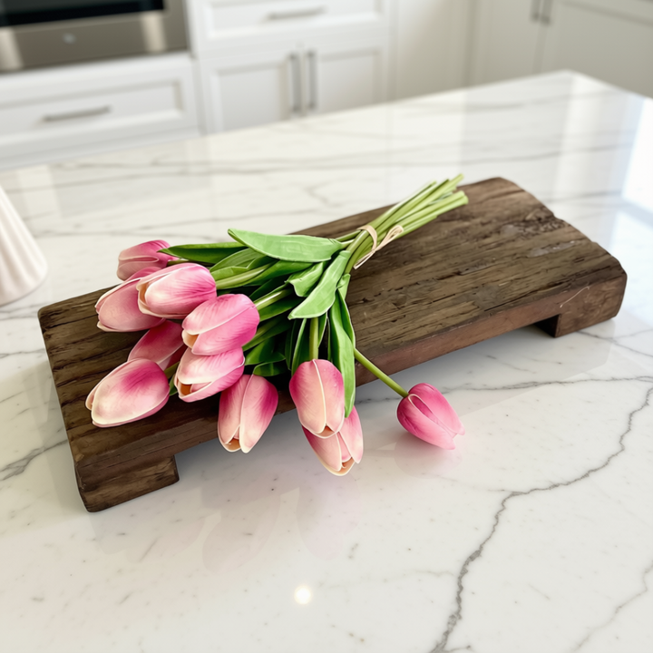 A bouquet of pink tulips with green stems rests on a rustic dark wood decorative riser placed on a white marble kitchen countertop.