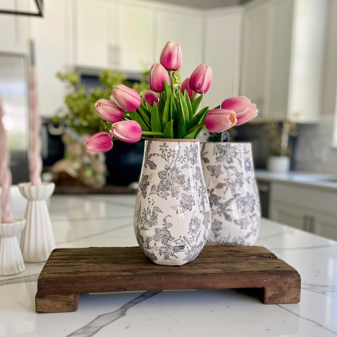 A white ceramic vase with a gray floral toile pattern holds pink tulips, styled on a dark found wooden riser on a white marble kitchen counter with a blurred kitchen background.