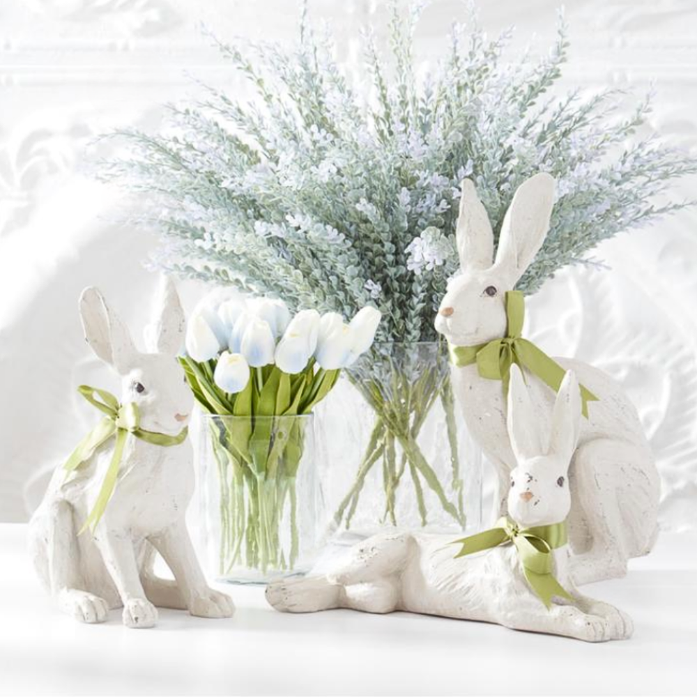 White rabbit figurines with green ribbons in front of a vase with blue tulips on a light background