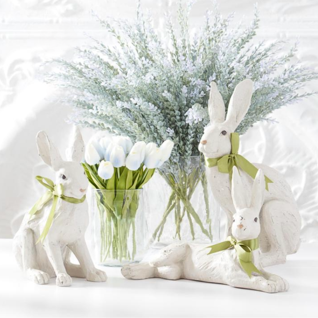 White rabbit figurines with green ribbons in front of a vase with blue tulips on a light background