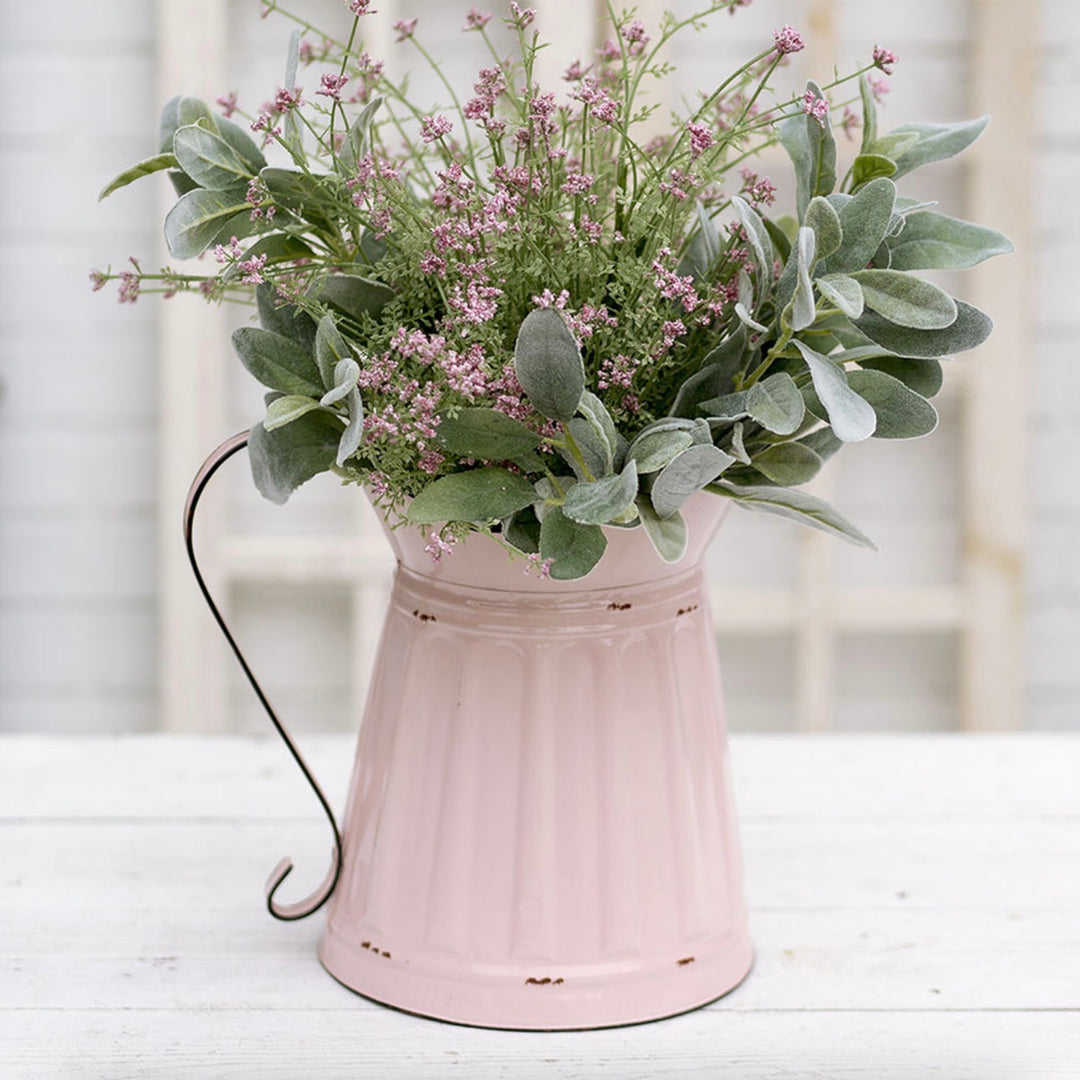 Pink metal vintage style wide mouth pitcher with greenery and small pink flowers on a white surface.