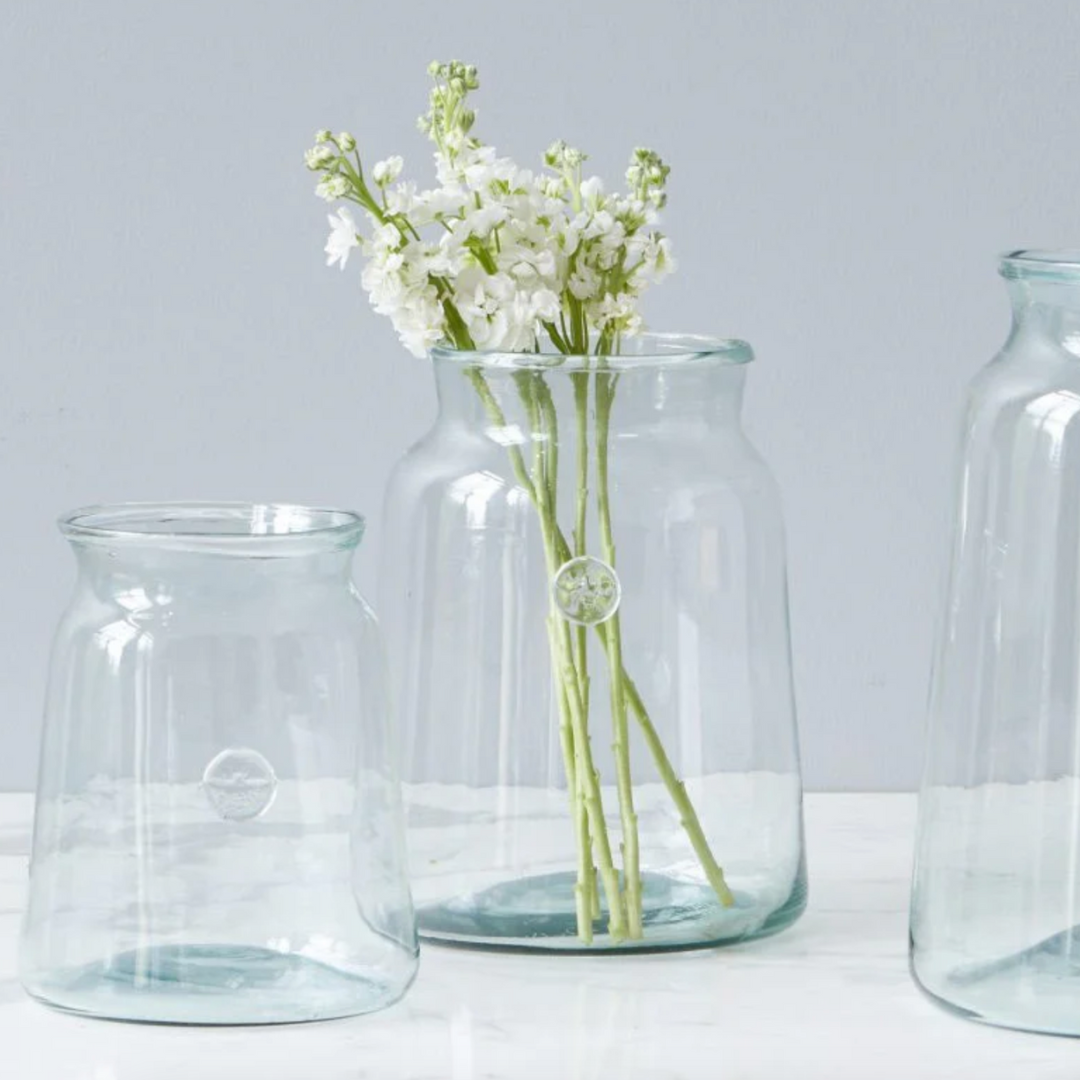 A clear glass French mason jar with a vintage-inspired bee seal, containing white flowers, placed on a white surface next to two other jars.