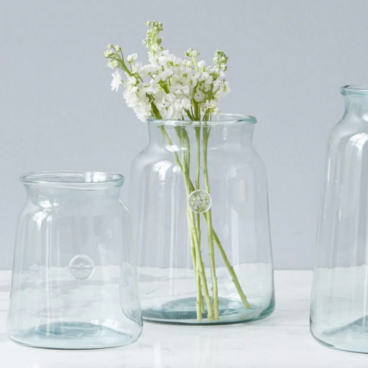 A clear glass French mason jar with a vintage-inspired bee seal, containing white flowers, placed on a white surface next to two other jars.