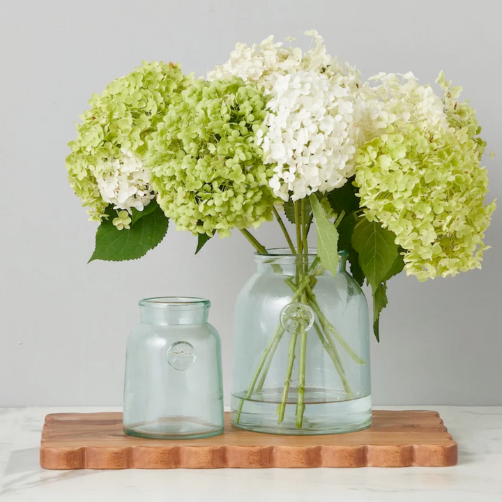 Bouquet of green and white hydrangeas in a clear glass French mason jar on a wooden board with a light gray background.