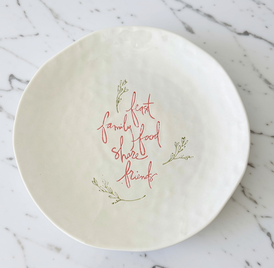 An off-white stoneware platter with a hand-painted red cursive quote reading 'feast family food share friends' and small green botanical illustrations, set on a white marble surface.