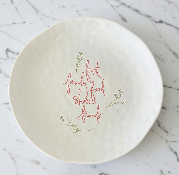 An off-white stoneware platter with a hand-painted red cursive quote reading 'feast family food share friends' and small green botanical illustrations, set on a white marble surface.