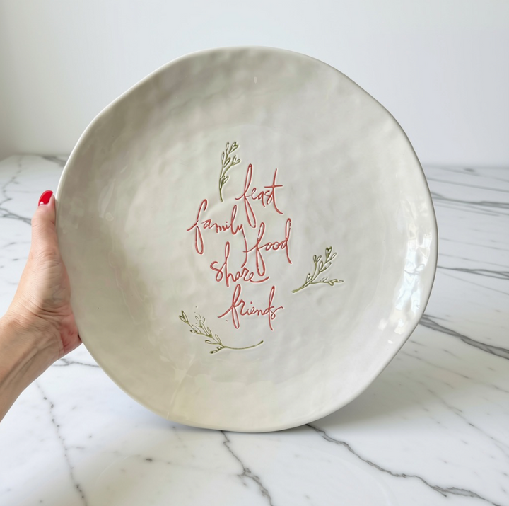 A hand with red nail polish holds a large, round, textured stoneware platter. The plate is off-white with a hand-painted script in the center reading 'family feast food share friends' in a reddish-brown color, accented with small green leaf illustrations.