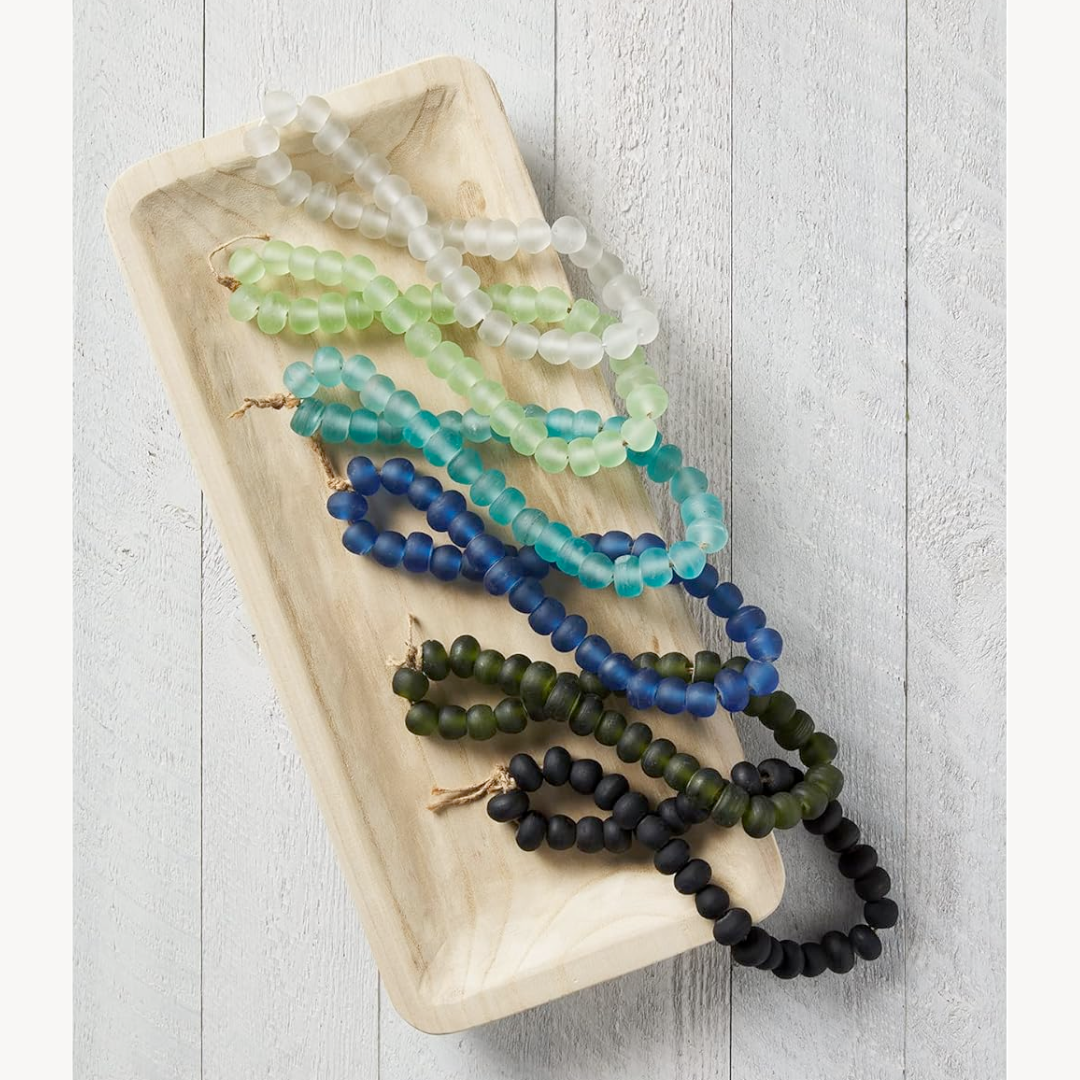 Strands of glass beads in various shades of blue and green, laid out on a wooden dish.